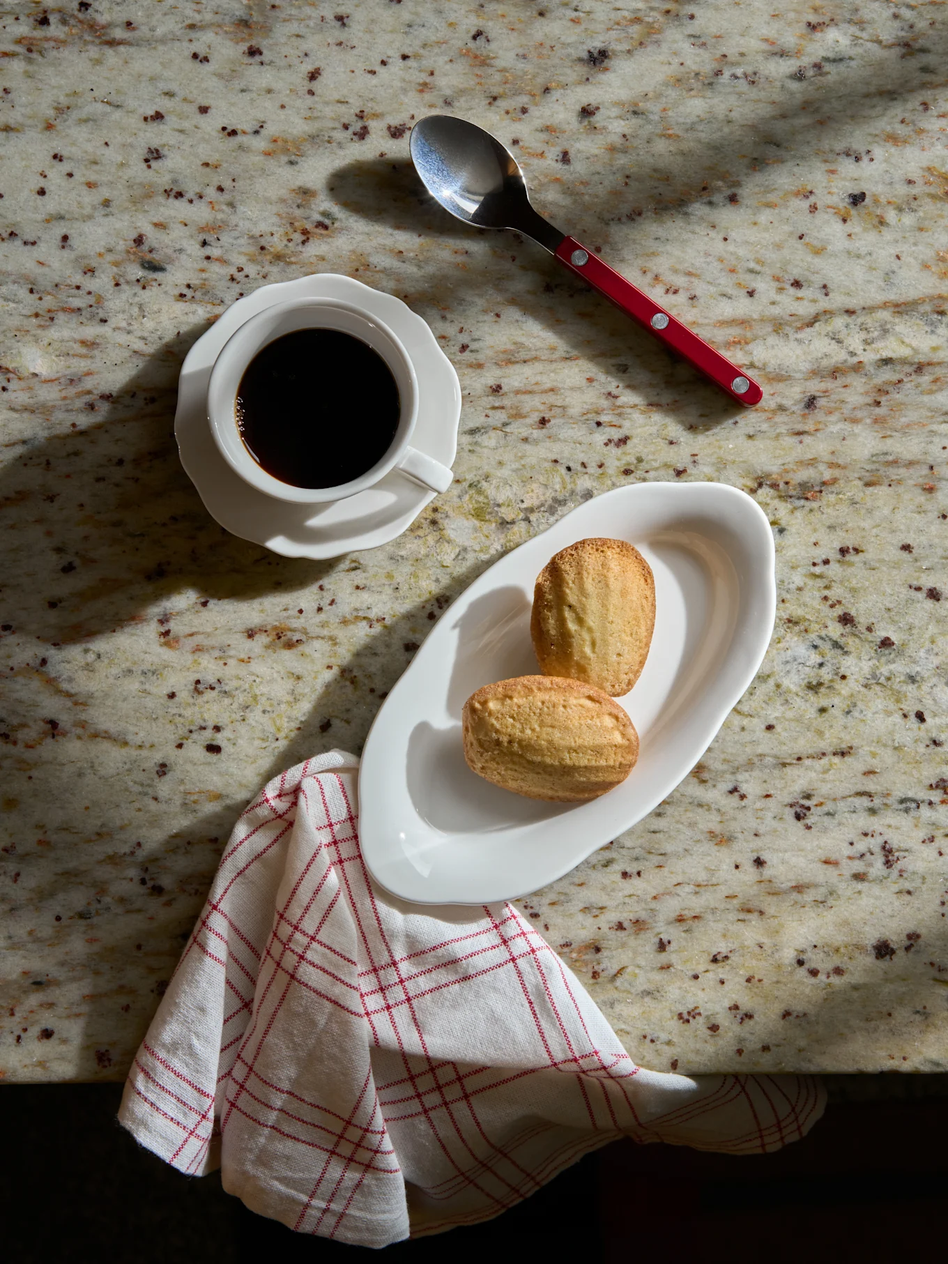Vista dall'alto: caffè nero in tazza bianca, due madeleines su piatto, cucchiaio e tovagliolo a righe su bancone in marmo.