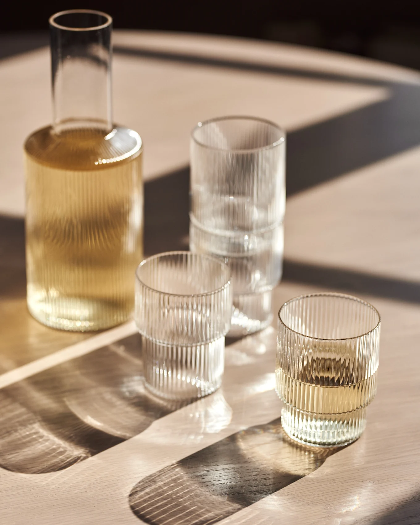 Caraffa di vetro e bicchieri a coste su un tavolo di legno con luce solare.