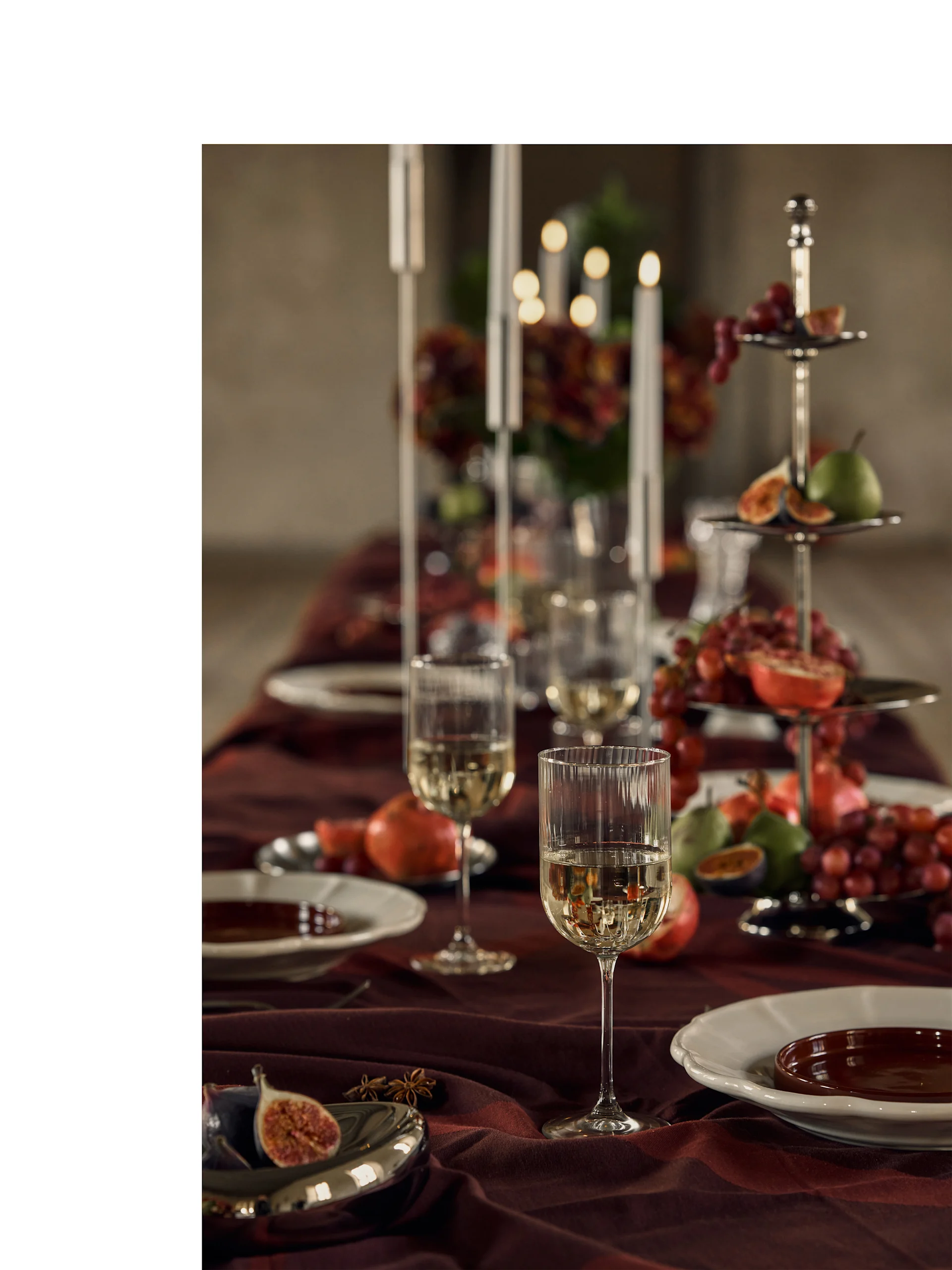 Tavola festiva apparecchiata con bicchieri di vino bianco, piatti e un'alzata a più piani con frutta colorata e candele.
