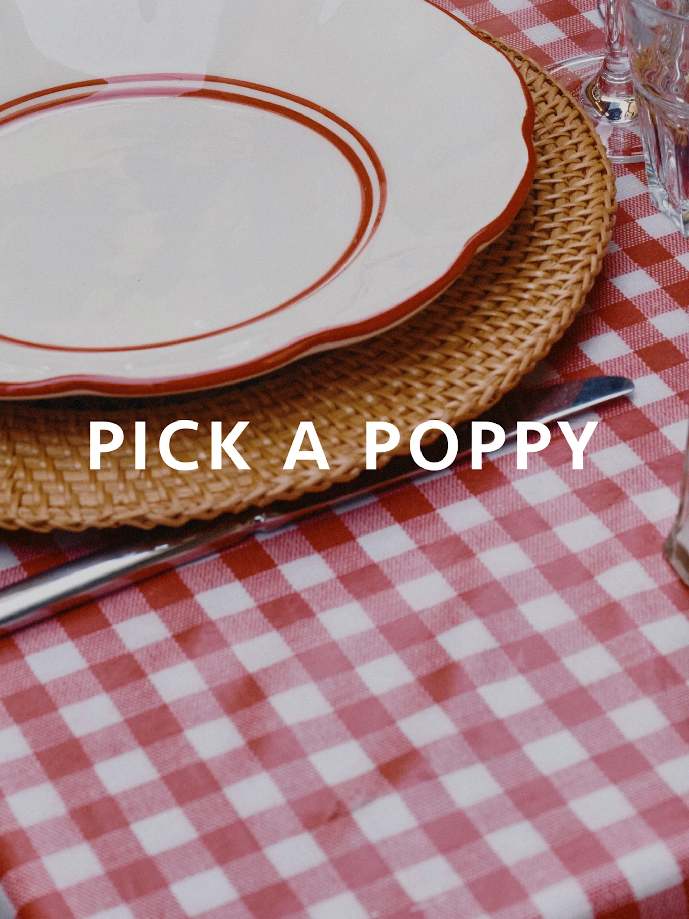 Tavola apparecchiata con tovaglia a quadri rossi e bianchi, tovaglietta in vimini e piatto bianco con bordo rosso. Testo: PICK A POPPY.