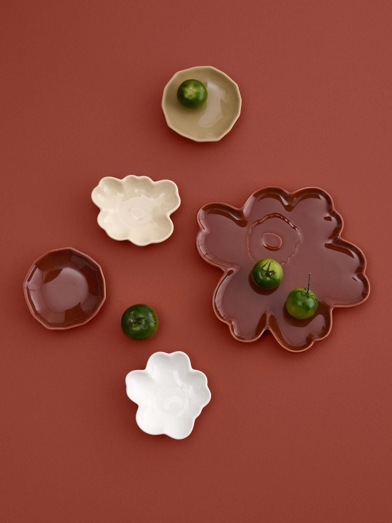 Assortimento di piccoli piatti dalle forme irregolari in crema, bianco e marrone con piccoli frutti verdi, su sfondo marrone-rossiccio.