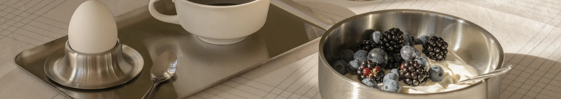 Vassoio da colazione con uovo sodo, caffè e una ciotola di yogurt con mirtilli e more su una tovaglia a quadri.