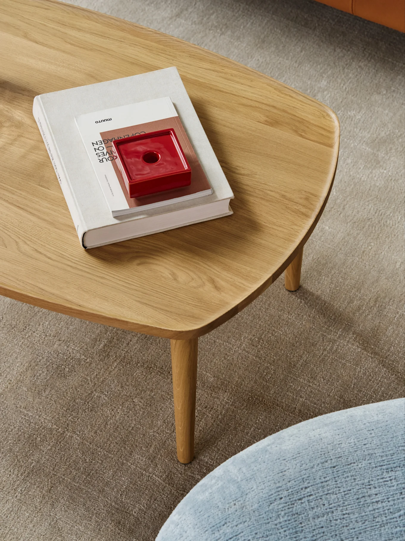 Un tavolino da caffè in legno chiaro con bordi arrotondati, con una pila di libri e un oggetto in ceramica rosso, su un tappeto beige.