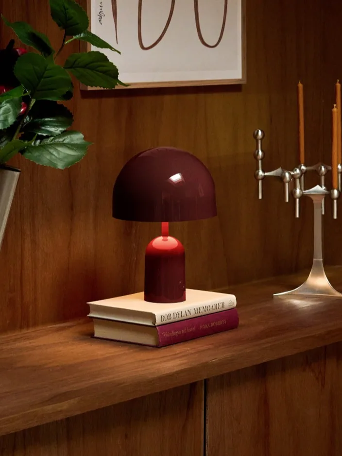 Mensola in legno: vaso scanalato argento con fiori rosso scuro, lampada bordeaux accesa su libri e candelabro argento.