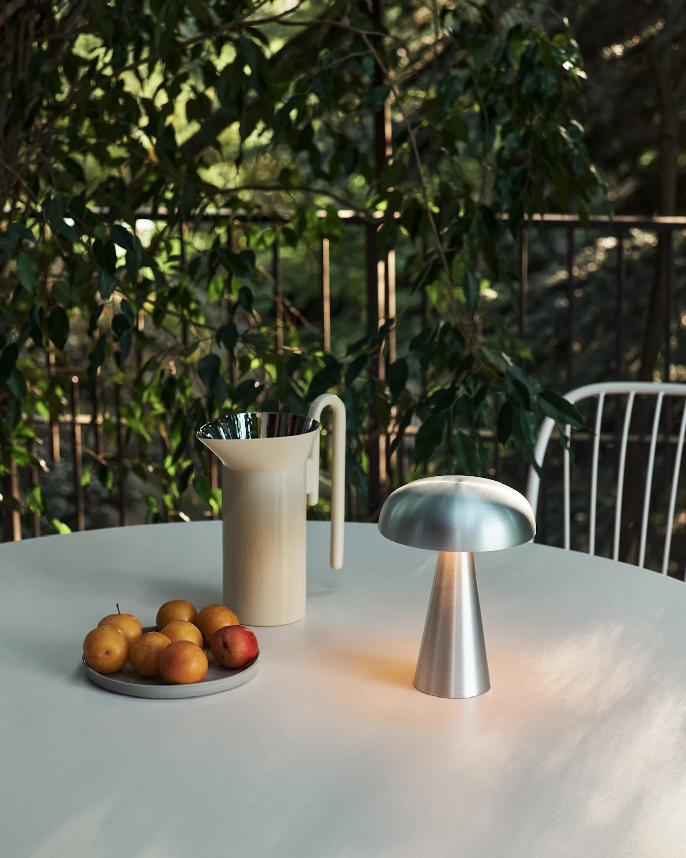 Tavolo da esterno con lampada a fungo argentata, brocca color crema e prugne tra il fogliame verde.