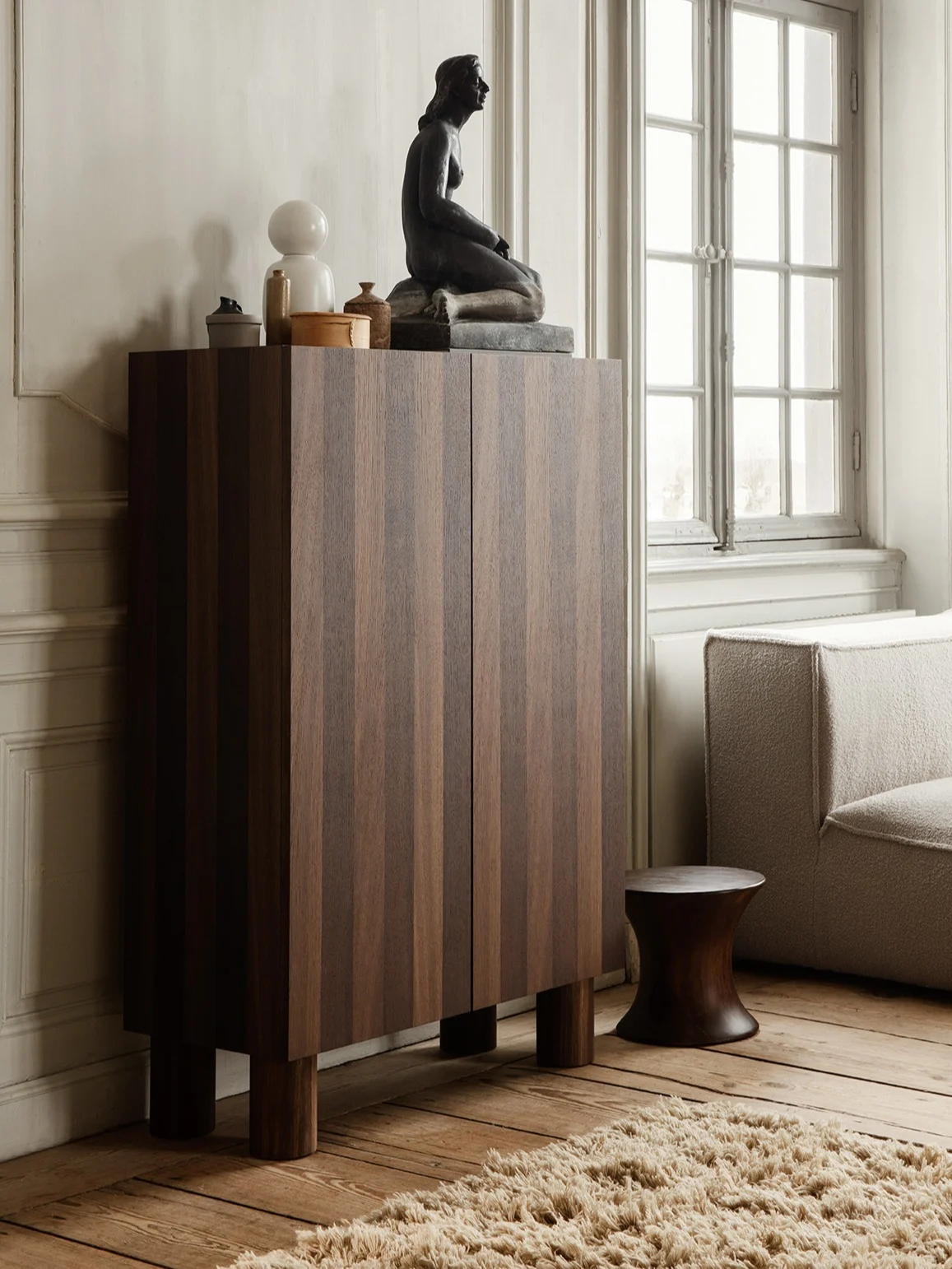 Armadio in legno scuro a righe verticali, sormontato da una scultura seduta, lampada bianca e decorazioni, accanto a divano e sgabello scuro.