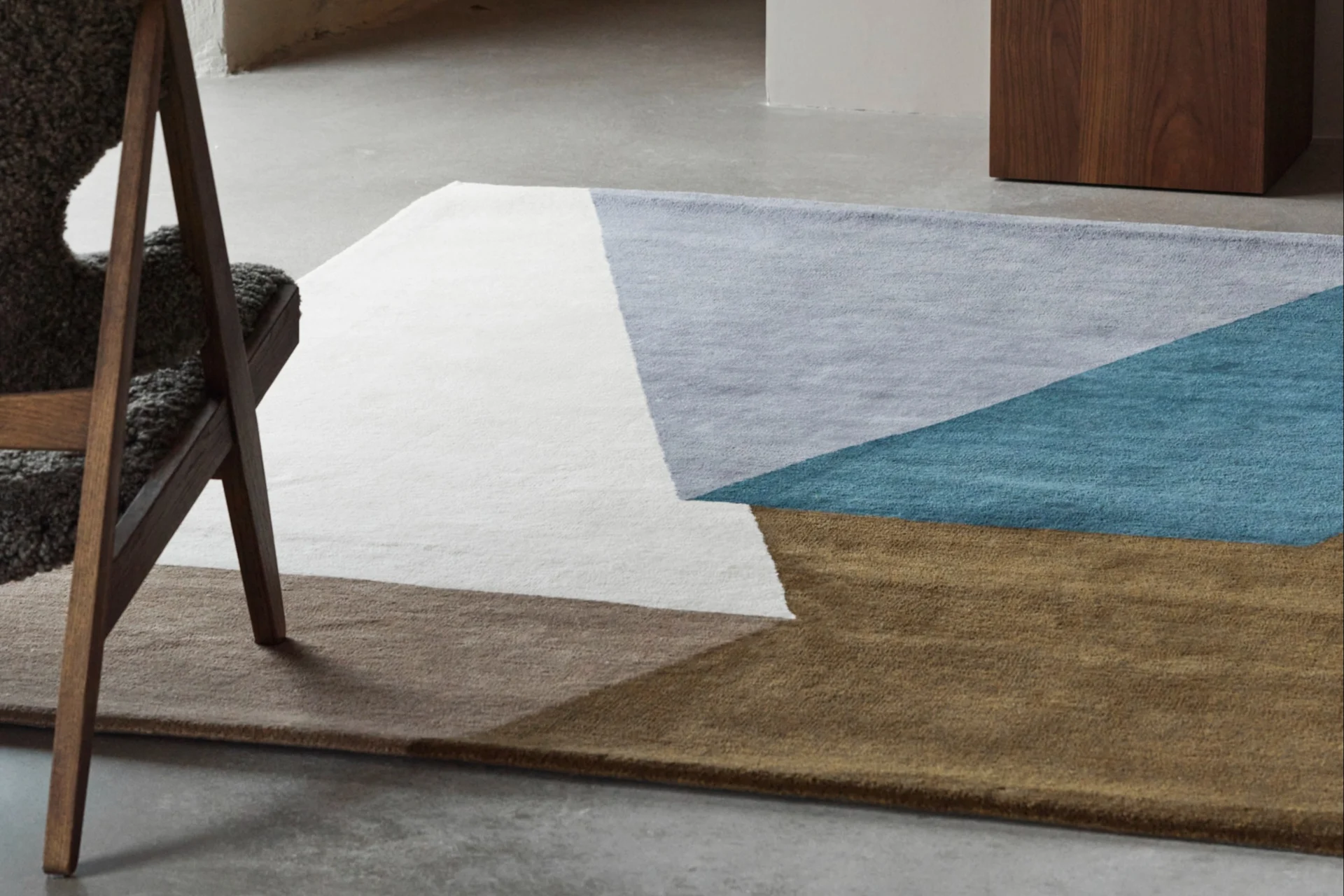 Un tappeto geometrico bianco, azzurro, turchese e marrone su un pavimento in cemento, con una sedia di legno e un vaso blu.