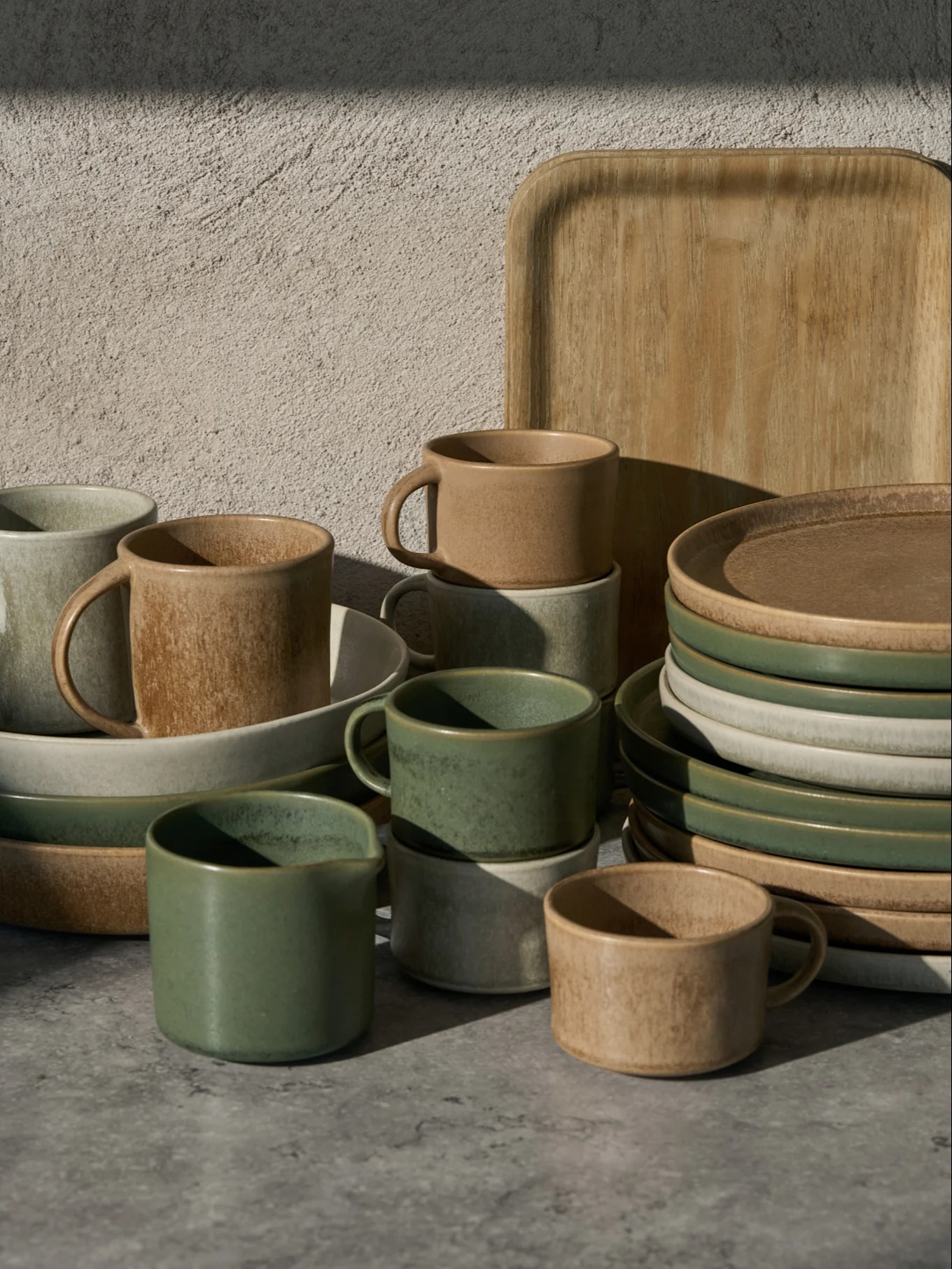 Collezione di tazze, ciotole e piatti in ceramica verdi, beige e grigio chiaro, con un vassoio di legno, su una superficie grigia.