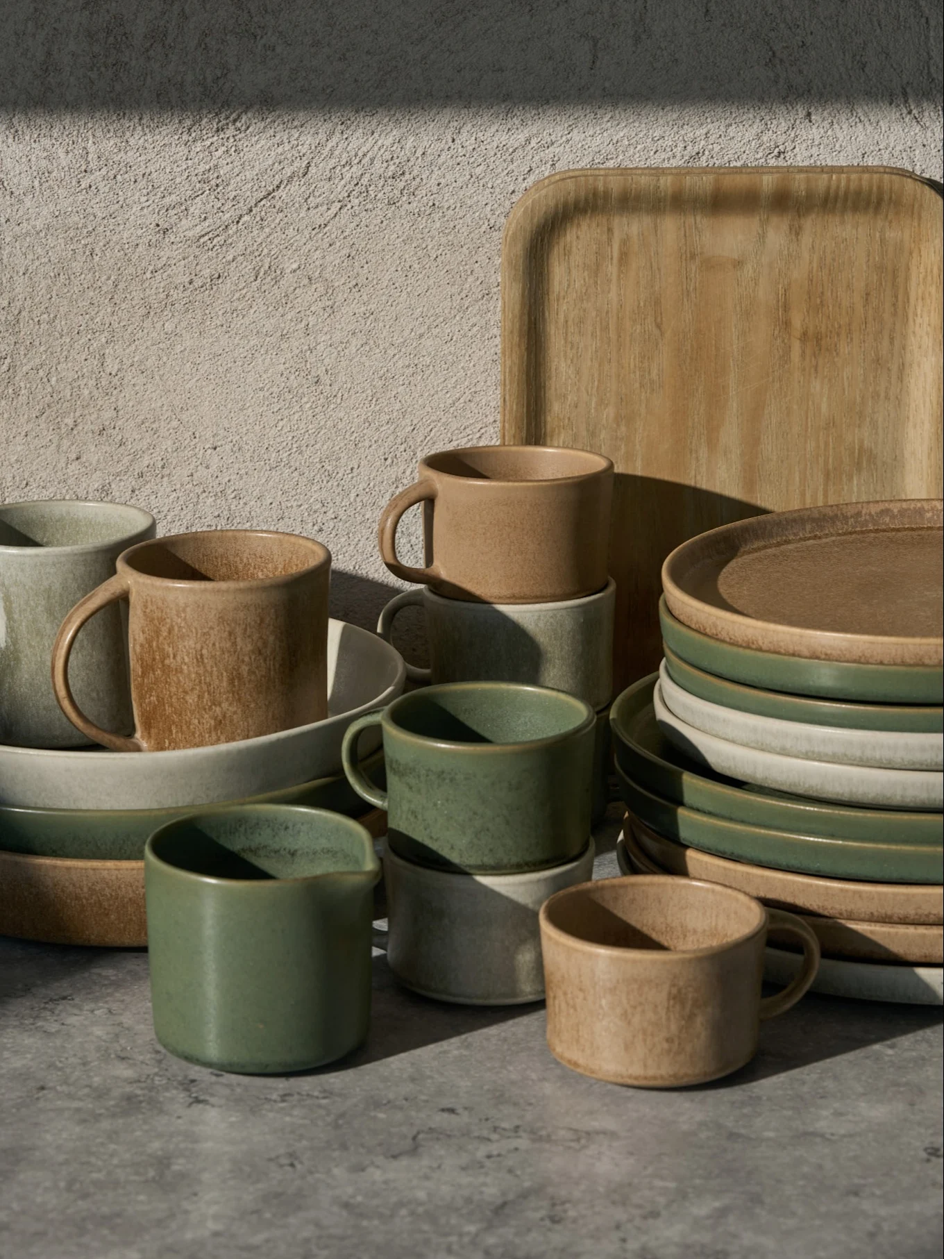 Collezione di tazze, ciotole e piatti in ceramica verdi, beige e grigio chiaro, con un vassoio di legno, su una superficie grigia.