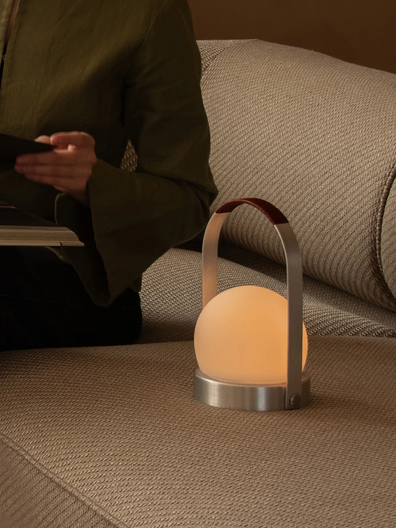 Persona che legge su un divano con una lampada portatile luminosa con globo satinato, base in metallo e manico in pelle.