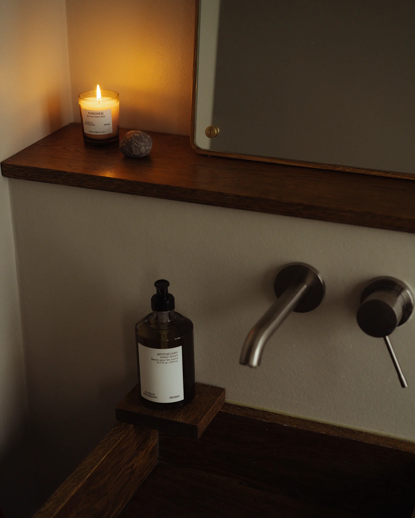 Bagno accogliente con candela accesa, flacone di sapone per le mani e rubinetto in metallo spazzolato su lavabo in legno scuro.