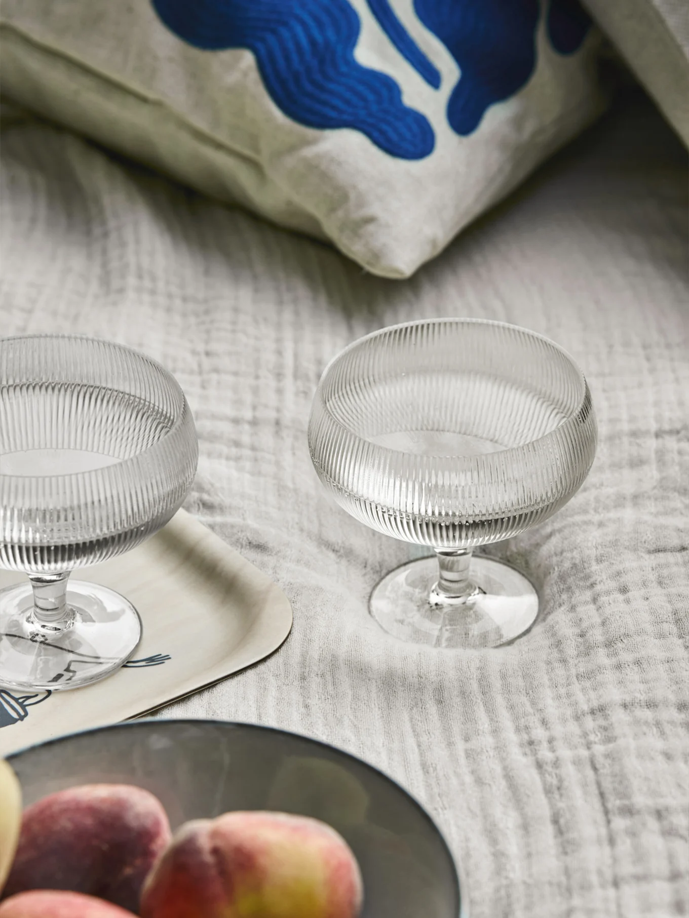 Due coppe da dessert in vetro a coste, un cuscino con arte astratta blu, un vassoio di legno e pesche su un tessuto chiaro.