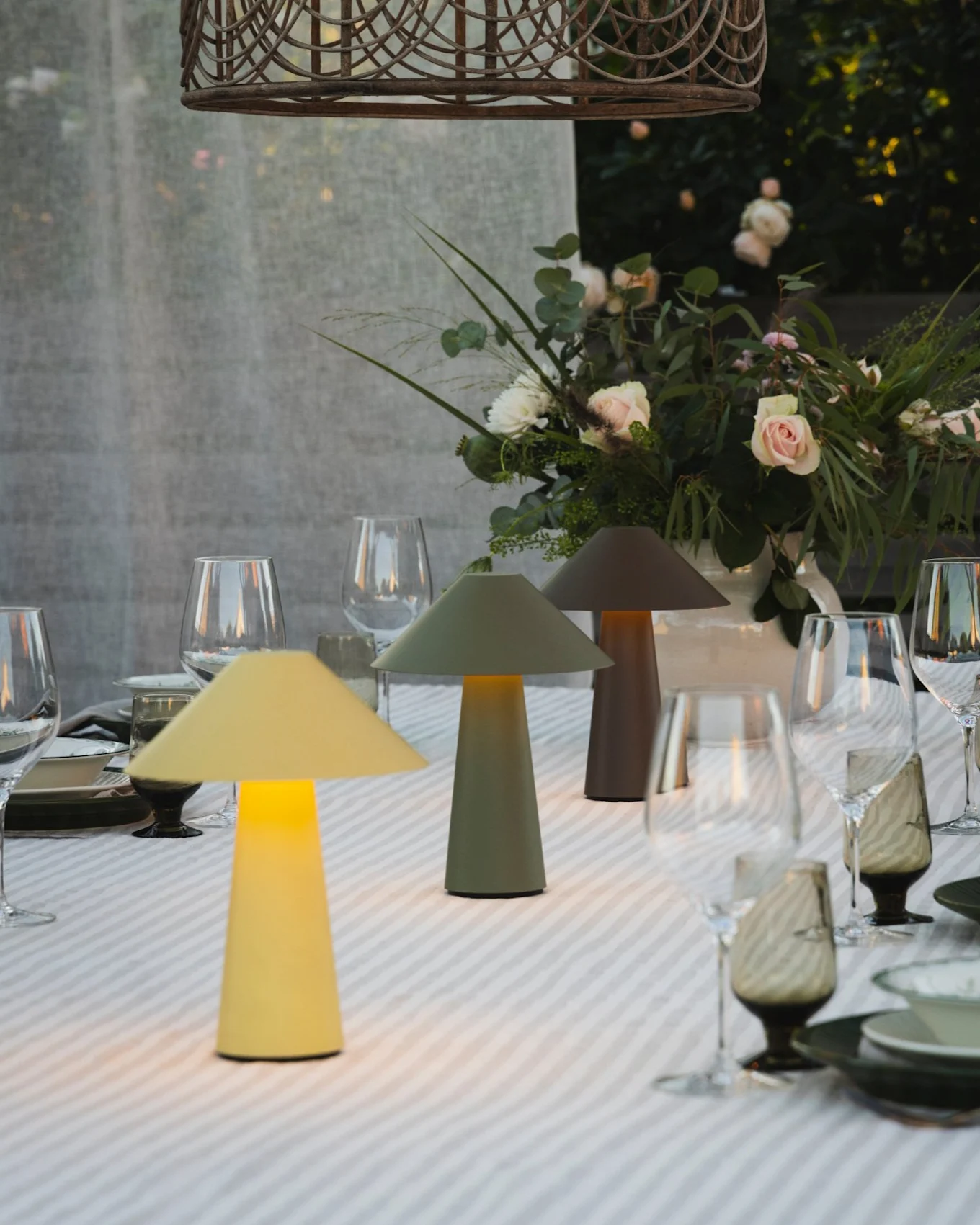 Tavolo da pranzo esterno con tre lampade a fungo illuminate, centrotavola floreale e lampada a sospensione in vimini.