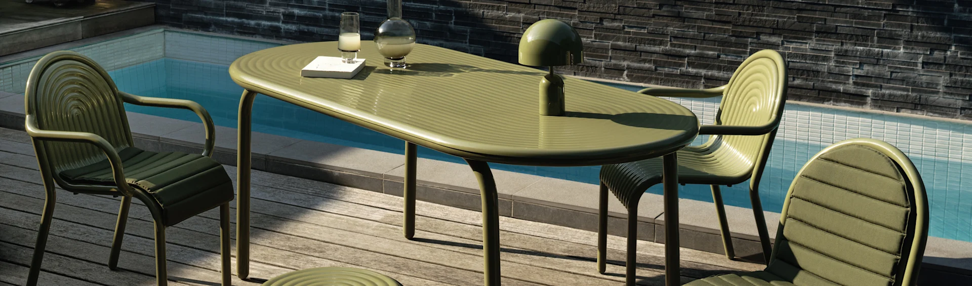 Tavolo e sedie da pranzo verdi a coste all'aperto su un ponte in legno a bordo piscina. Una lampada, una caraffa, un bicchiere e un libro sono sul tavolo.