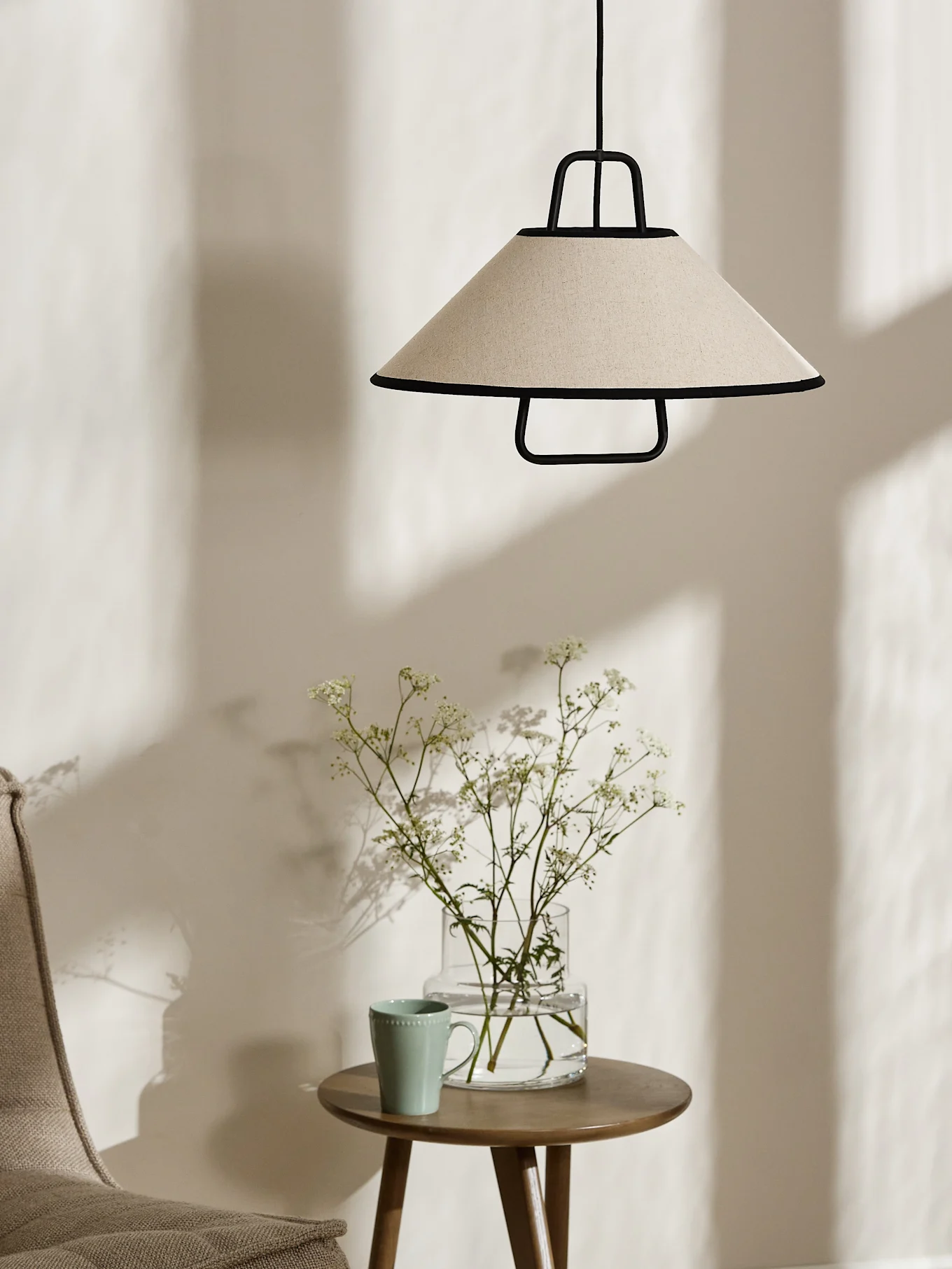 Una lampada a sospensione conica color crema con bordo nero pende sopra un tavolino in legno con un vaso di fiori bianchi e una tazza verde chiaro.
