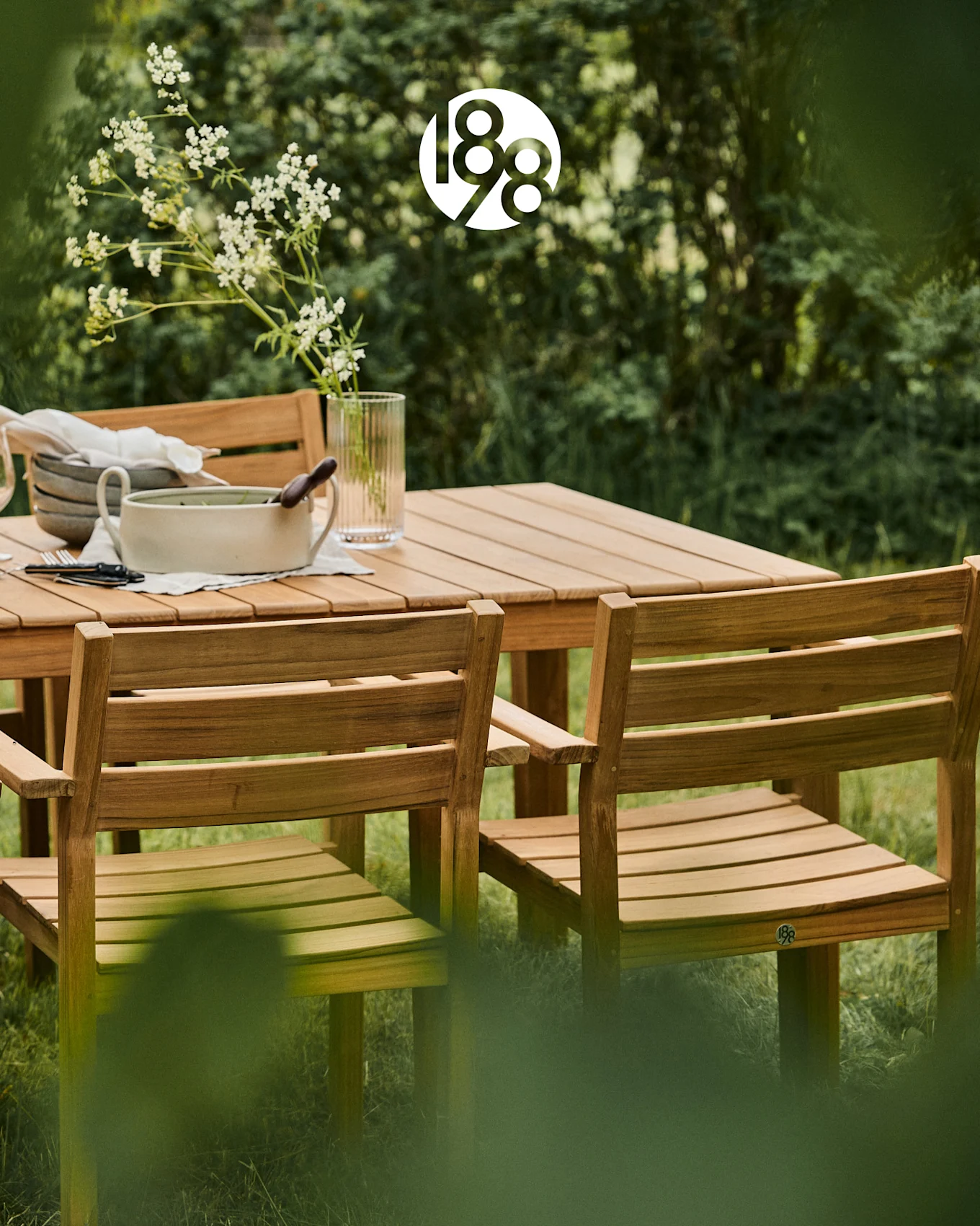 Set da pranzo esterno in legno con tavolo e sedie in un lussureggiante giardino verde, con fiori bianchi e piatti da portata.