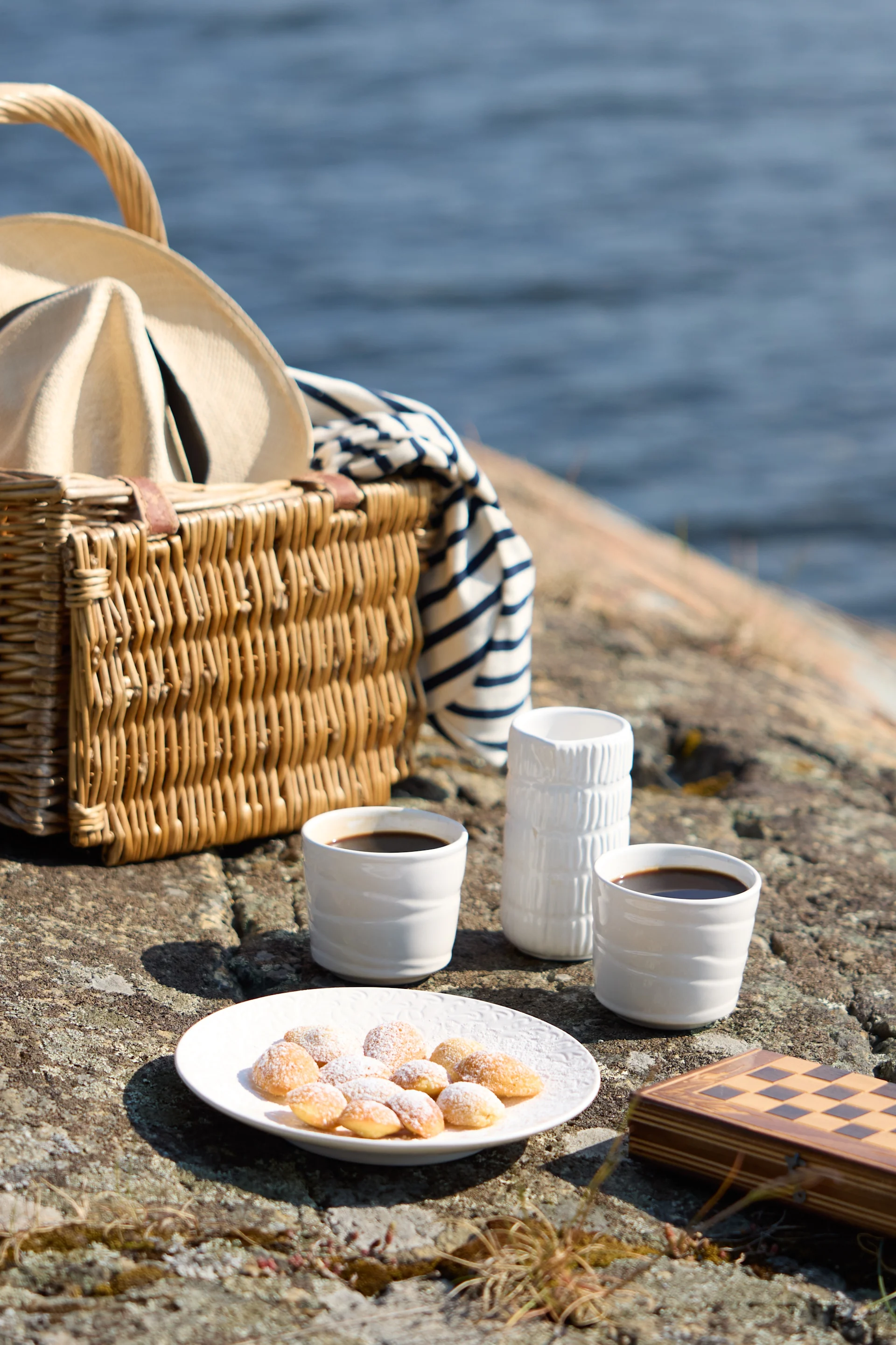 Un picnic su rocce in riva al mare, con un cestino di vimini con cappello e coperta a righe, due tazze di caffè, pasticcini e una scacchiera chiusa.