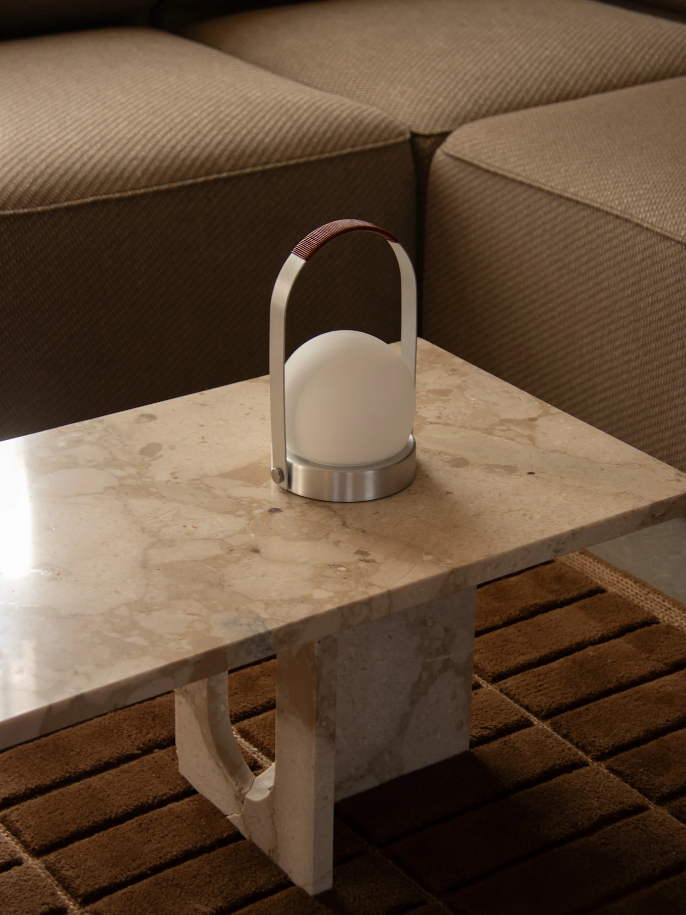 Lampada portatile moderna con luce bianca sferica, struttura argentata e manico in pelle marrone su un tavolo di marmo accanto a un divano.