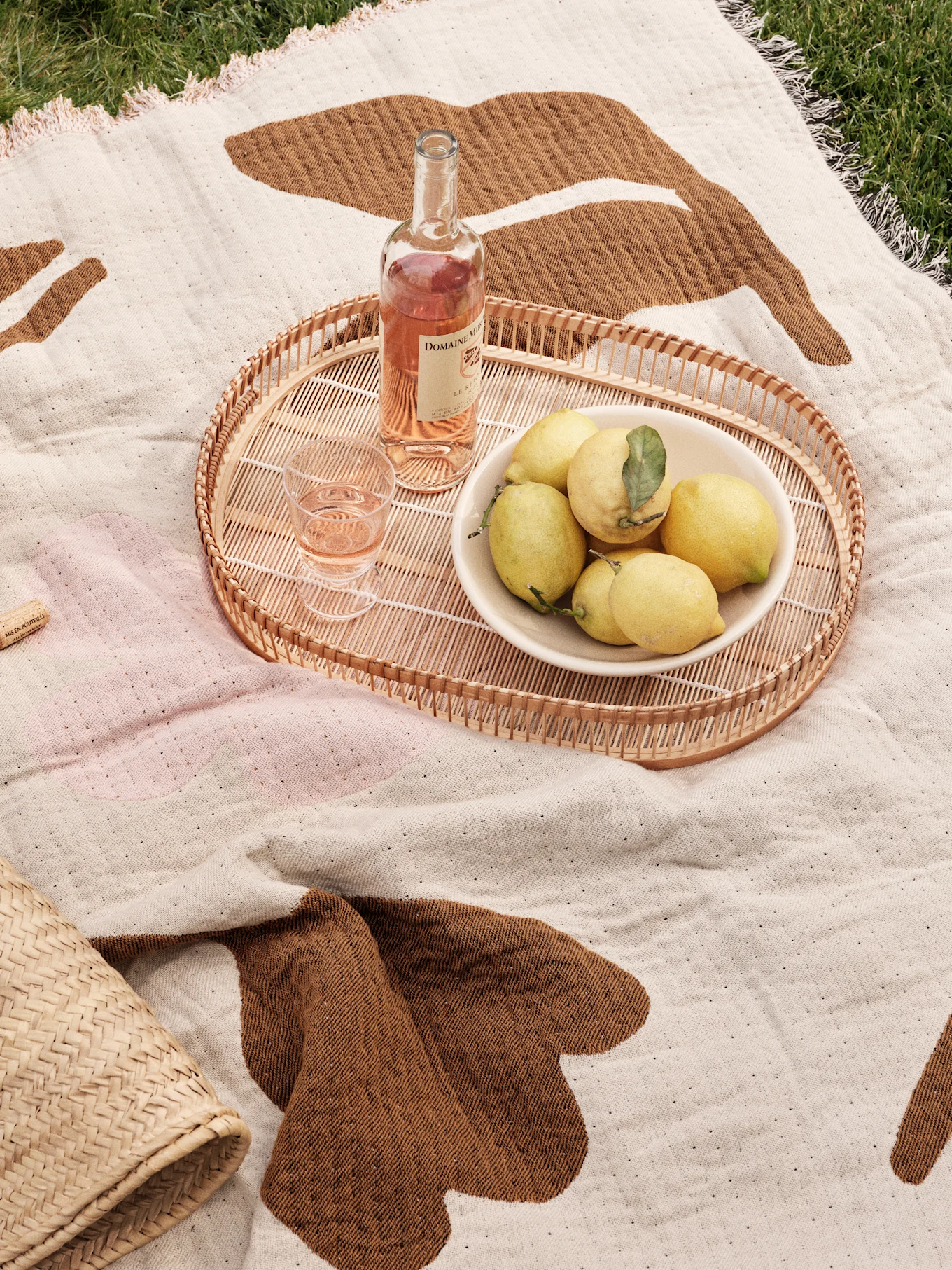 Coperta da picnic sull'erba con vassoio in rattan, vino rosé, bicchiere e una ciotola di limoni.