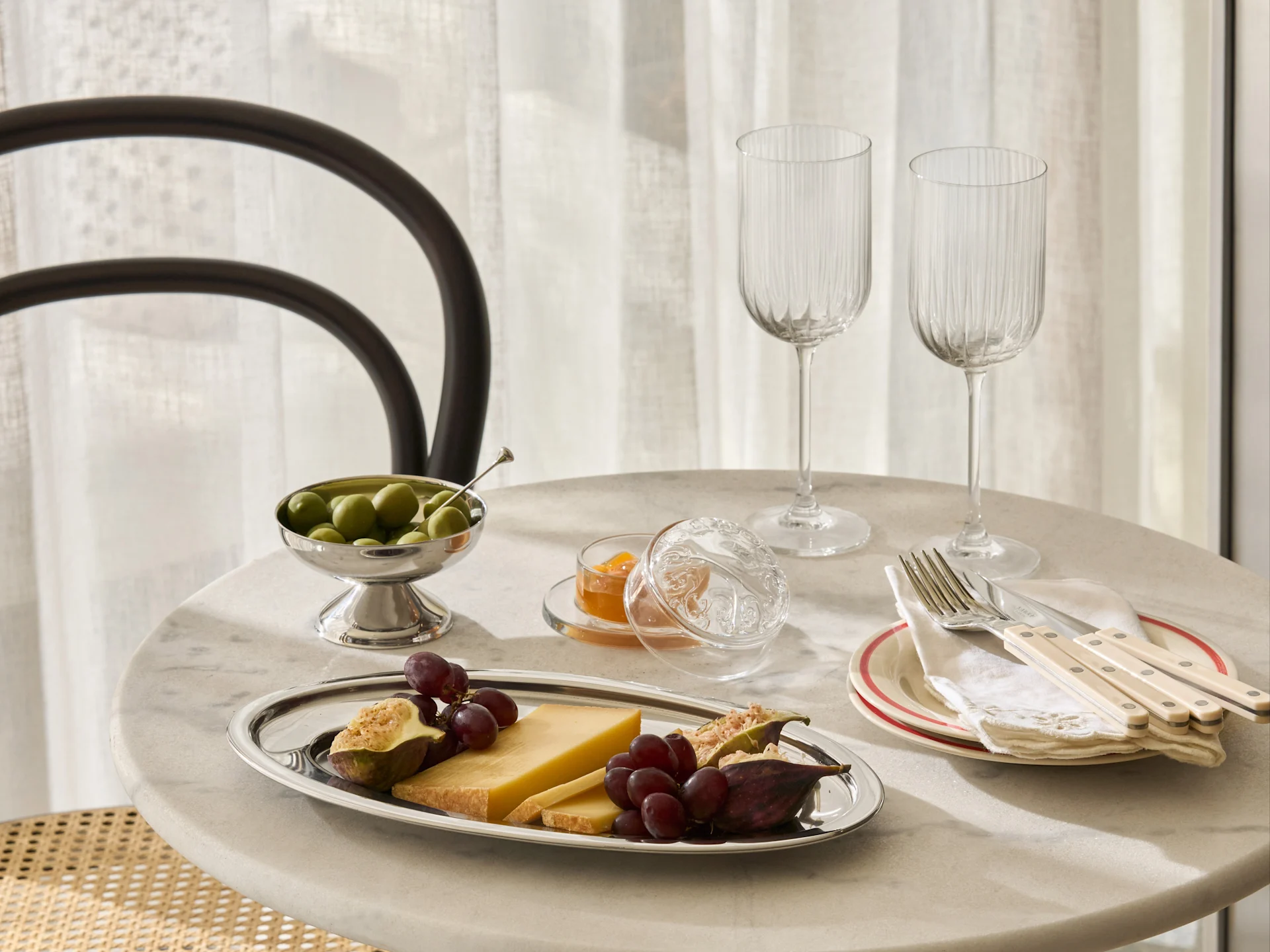 Un tavolo in marmo apparecchiato con un vassoio d'argento di formaggio, uva, fichi, una ciotola di olive, marmellata, due bicchieri a coste e posate sui piatti.