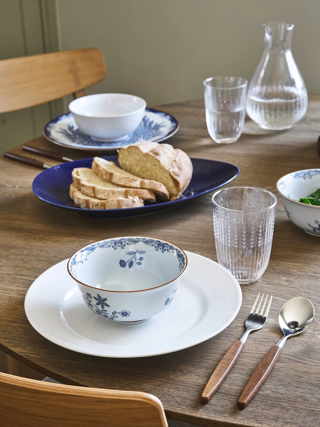 Tavolo da pranzo apparecchiato con due coperti, ciotole floreali blu e bianche, pane su un piatto blu e posate con manico in legno.