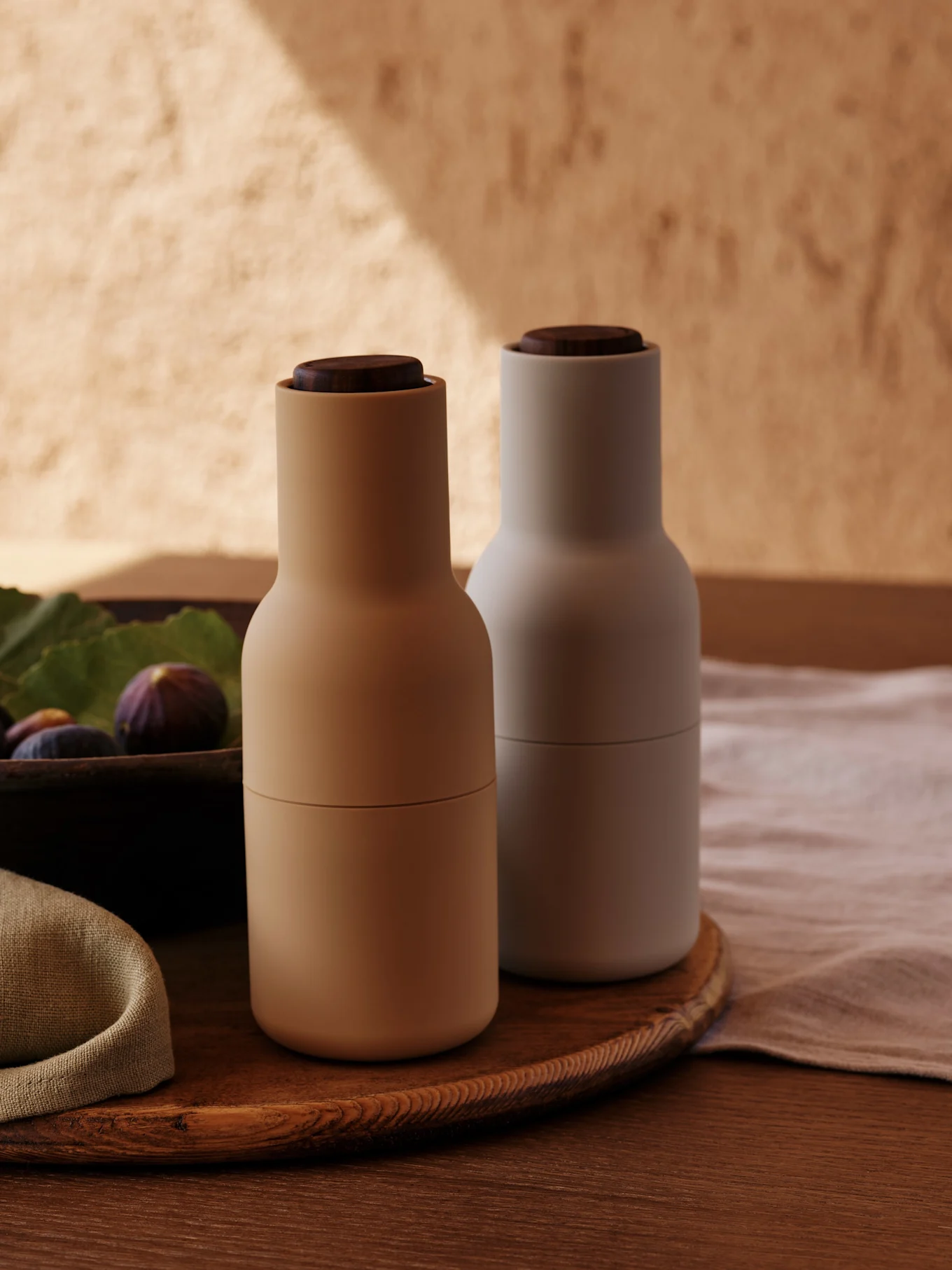 Due macinaspezie moderni con coperchi in legno, uno beige e uno grigio chiaro, su un vassoio di legno con fichi.