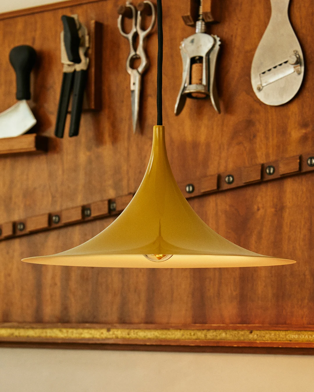 Lampada a sospensione dorata a forma di campana con lampadina visibile, su una parete in legno con attrezzi.