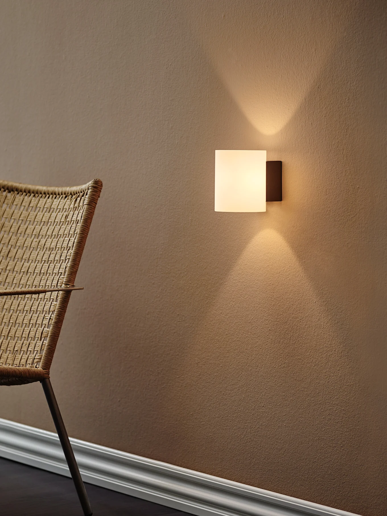 Moderna applique quadrata che proietta luce calda verso l'alto e il basso su una parete marrone testurizzata, con una sedia intrecciata a sinistra.