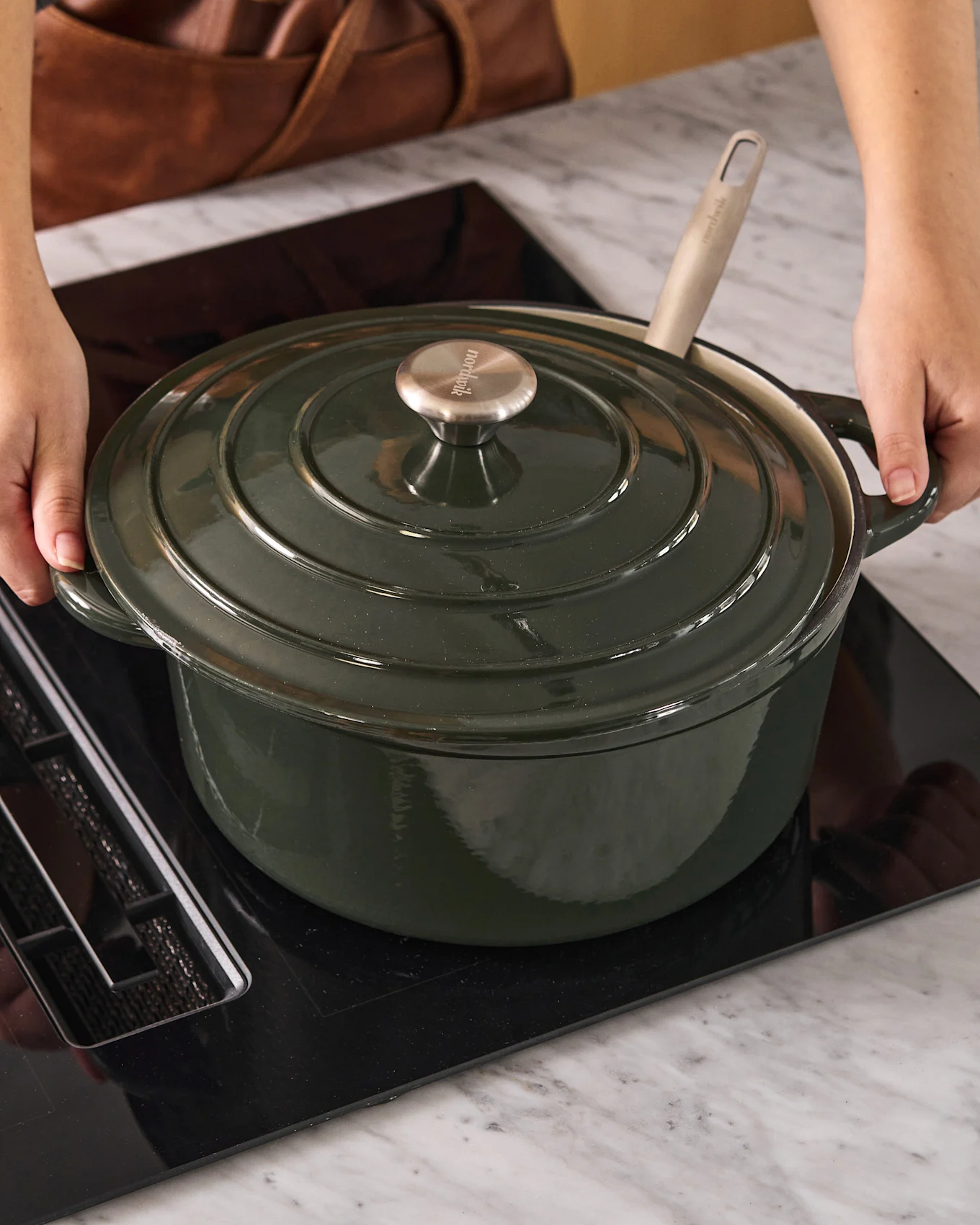 Persona che posiziona una casseruola olandese verde scuro con manopola in metallo su un piano cottura a induzione nero.