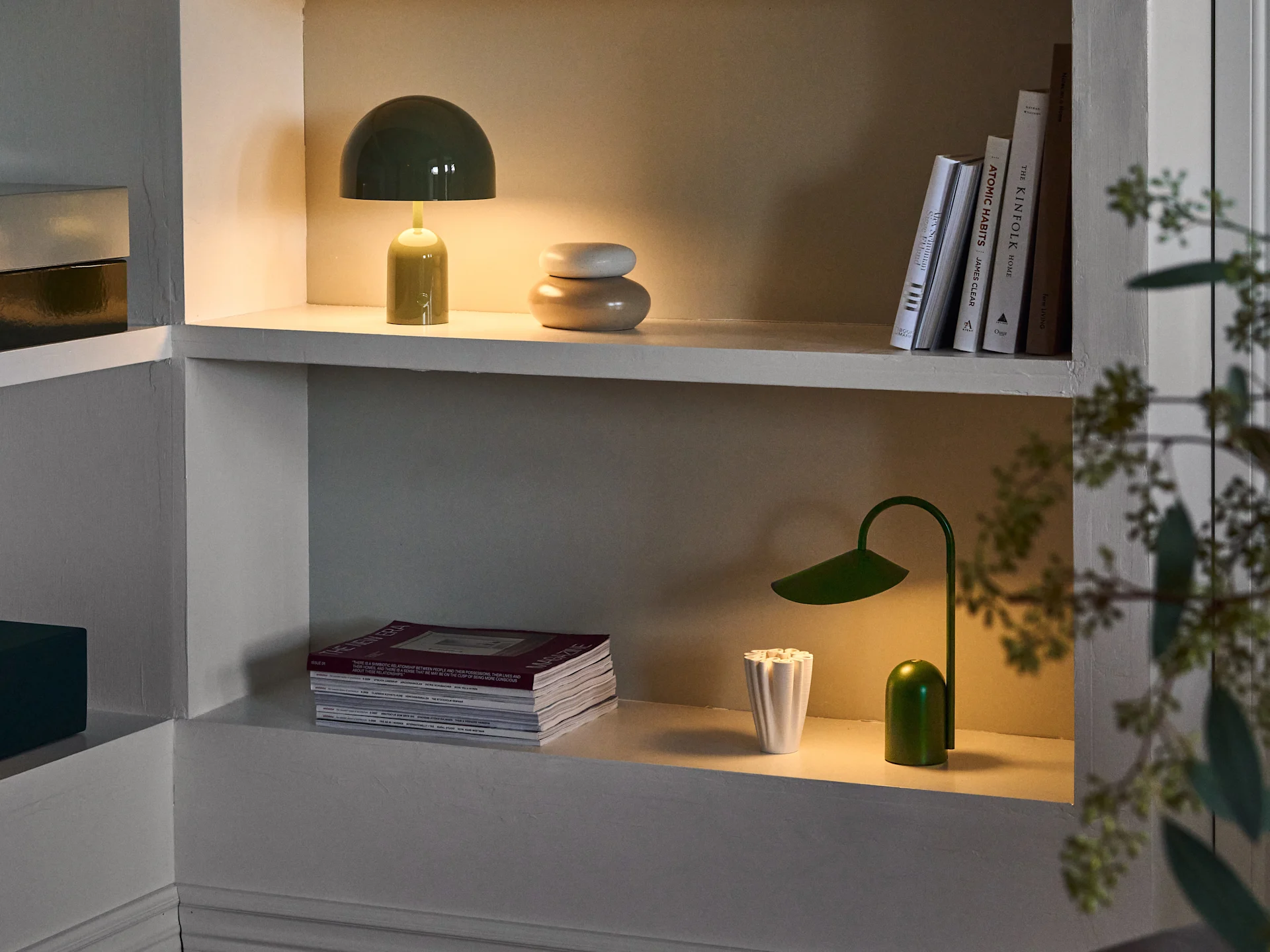 Mensole illuminate. Superiore: lampada a fungo verde, decorazioni beige impilate e libri. Inferiore: riviste, vaso bianco e lampada a foglia verde.