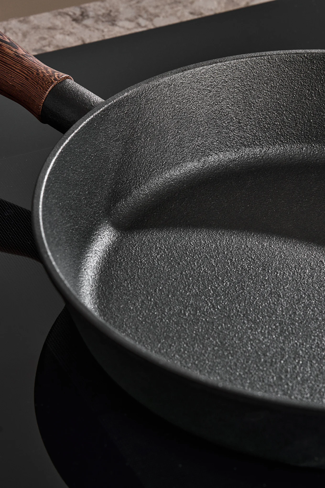 Primo piano di una padella scura e strutturata con manico in legno su un piano cottura nero.