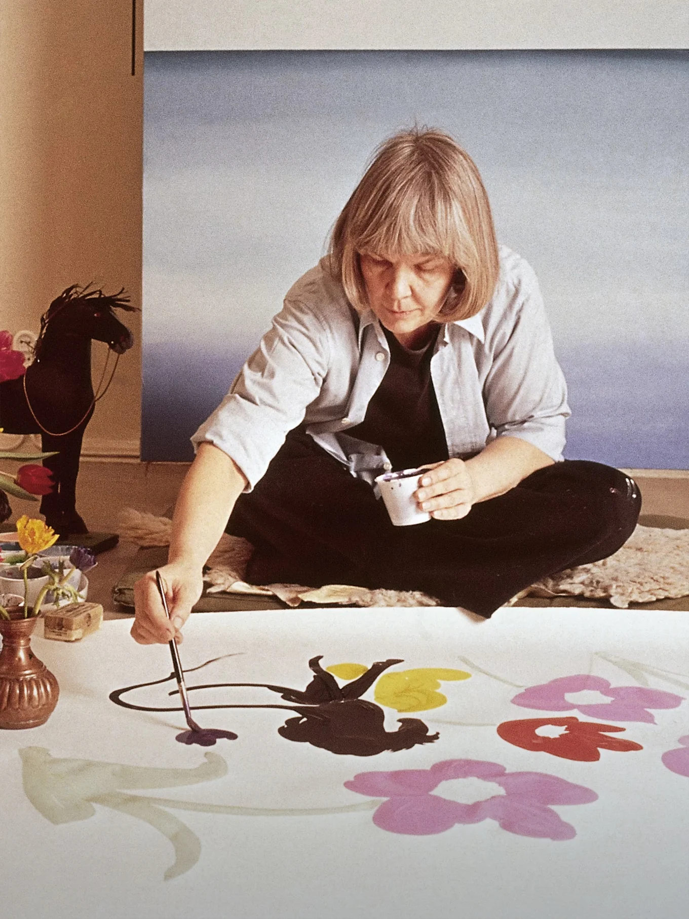 L'artista Marimekko Maija Isola seduta sul pavimento dipinge grandi fiori colorati su un tessuto bianco.