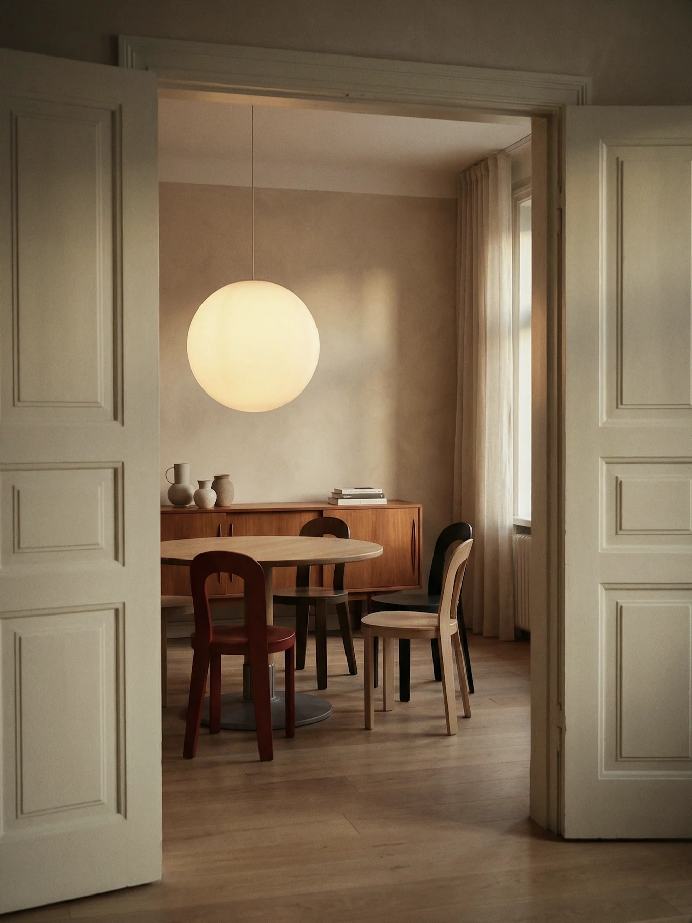 Sala da pranzo attraverso apertura, con tavolo rotondo in legno, sedie, grande lampada sferica e credenza.