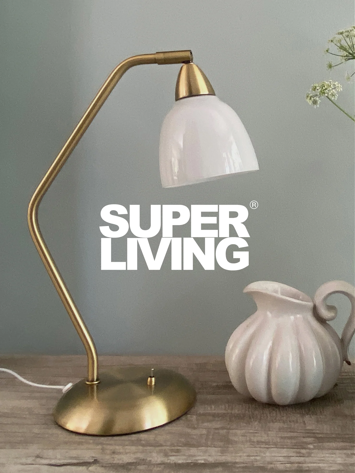 Marchio SUPER LIVING su una lampada da scrivania in ottone con paralume bianco e una brocca bianca scanalata su un tavolo di legno.