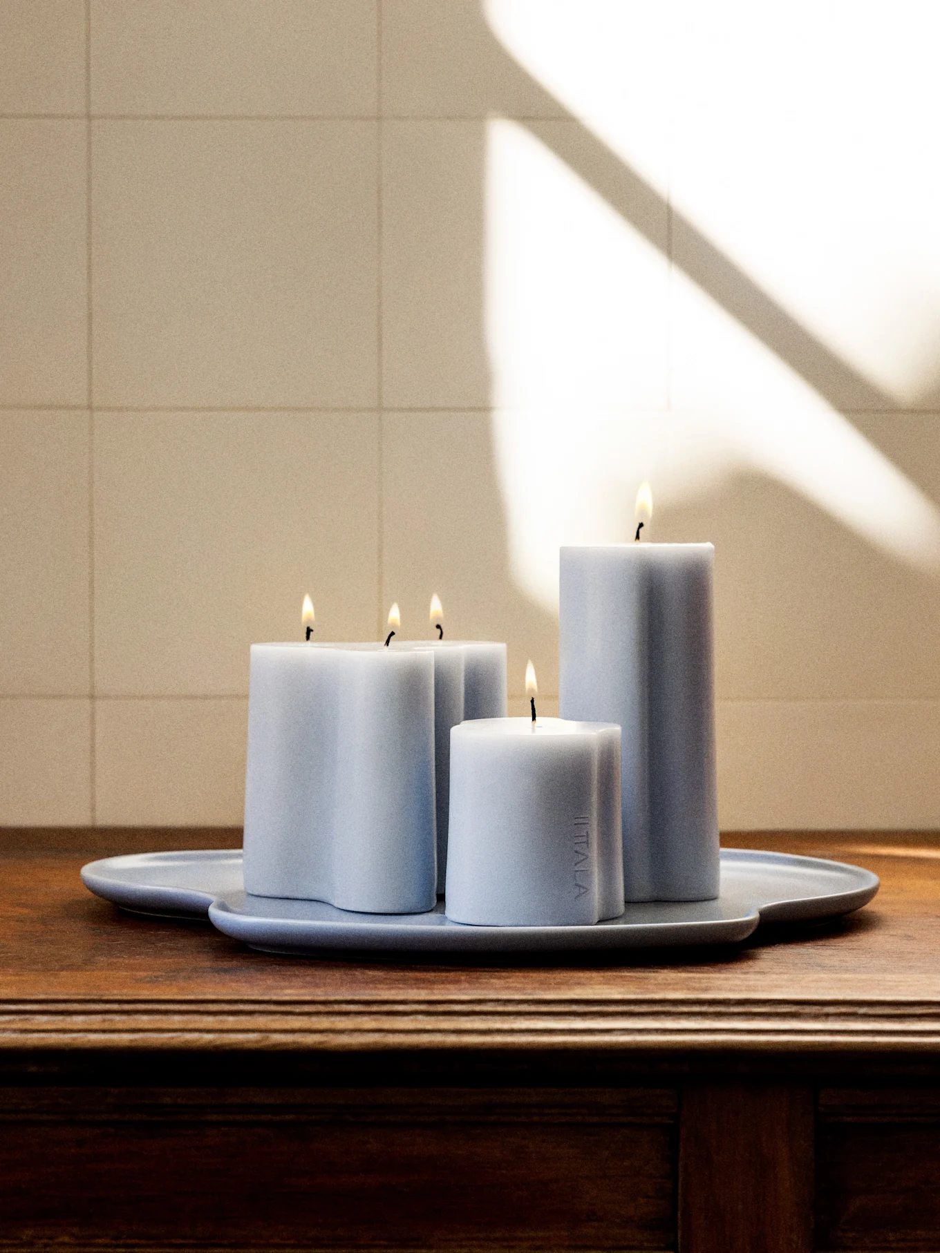 Quattro candele accese azzurro-grigie di varie forme e altezze su un vassoio abbinato di forma irregolare su una superficie di legno scuro.