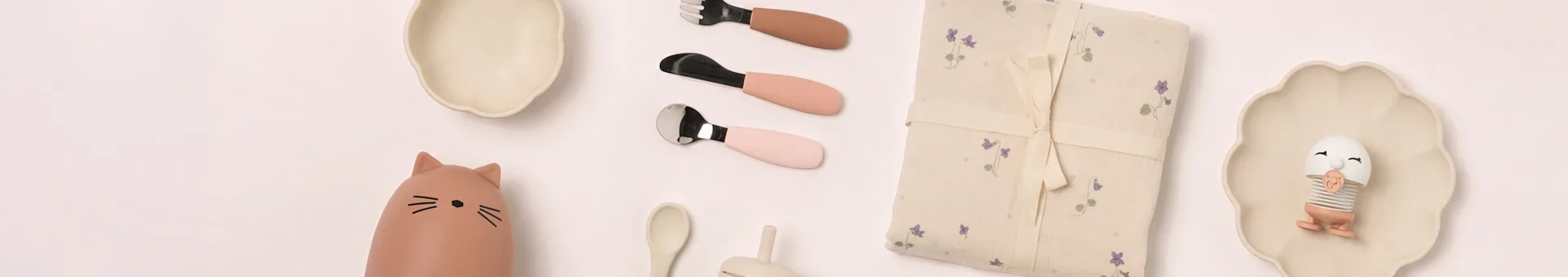 Articoli da pranzo per bambini a vista piatta: piatti beige, biberon gatto, posate rosa, coperta floreale, giocattolo a uovo.